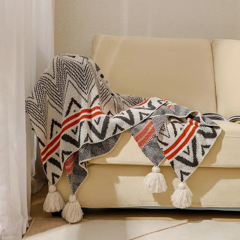 Bohemian style acrylic sofa blanket with tassel fringe in a geometric blue and pink pattern draped over a grey sofa.