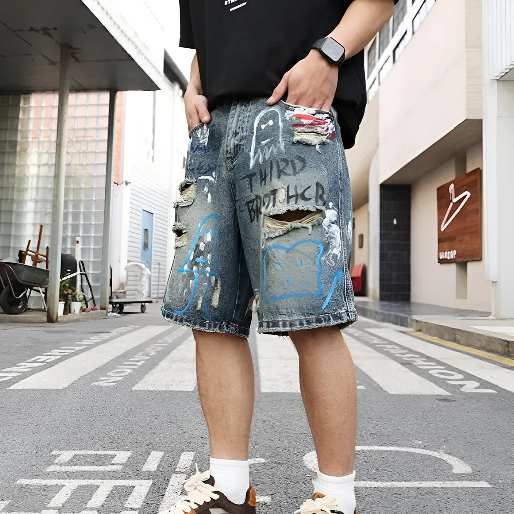 Blue men's ripped denim shorts with a graffiti pattern and zipper fly, laid flat.