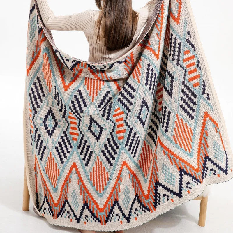 Bohemian style acrylic sofa blanket with tassel fringe in a geometric blue and pink pattern draped over a grey sofa.