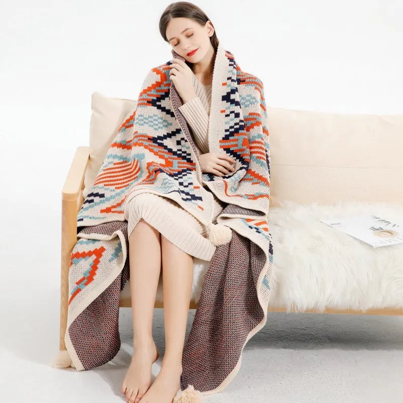 Bohemian style acrylic sofa blanket with tassel fringe in a geometric blue and pink pattern draped over a grey sofa.