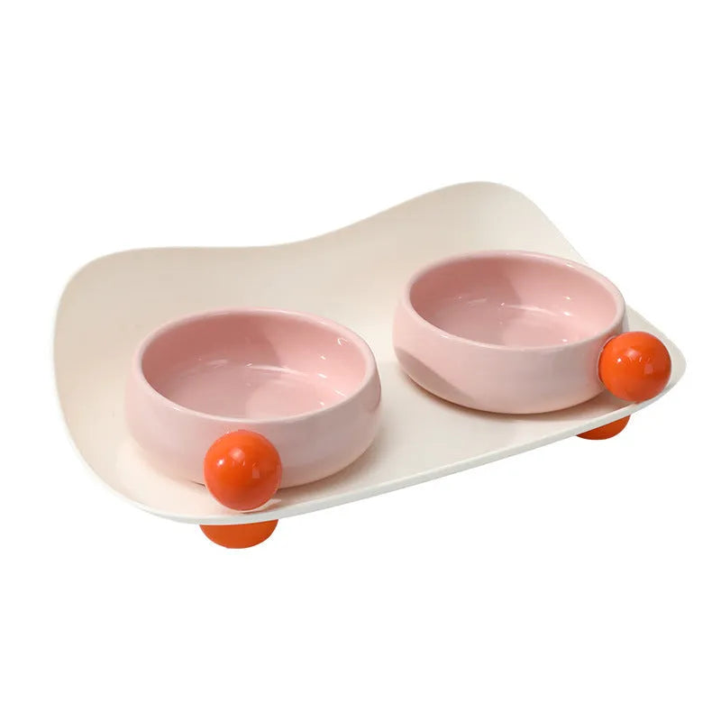 A pink ceramic raised pet bowl with two compartments and a non-slip black rubber base.