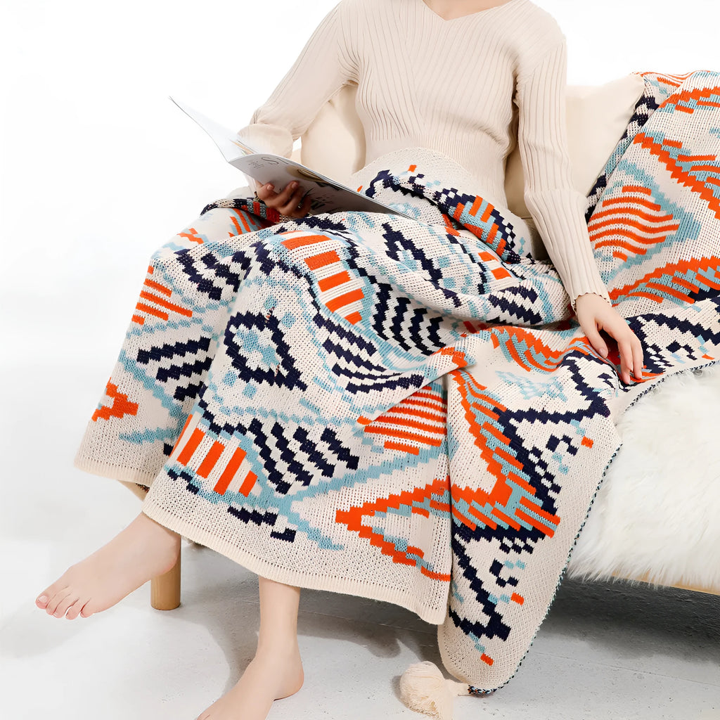 Bohemian style acrylic sofa blanket with tassel fringe in a geometric blue and pink pattern draped over a grey sofa.
