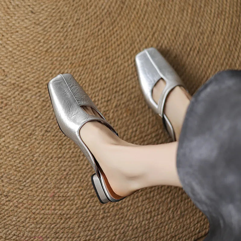 A pair of beige square head low heel slippers with a rubber sole on a wooden deck.