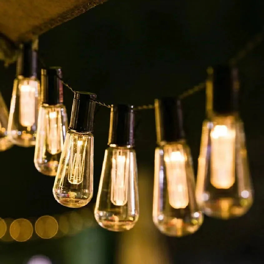 Retro glass infusion bottle-shaped outdoor string lights draped in a garden setting.