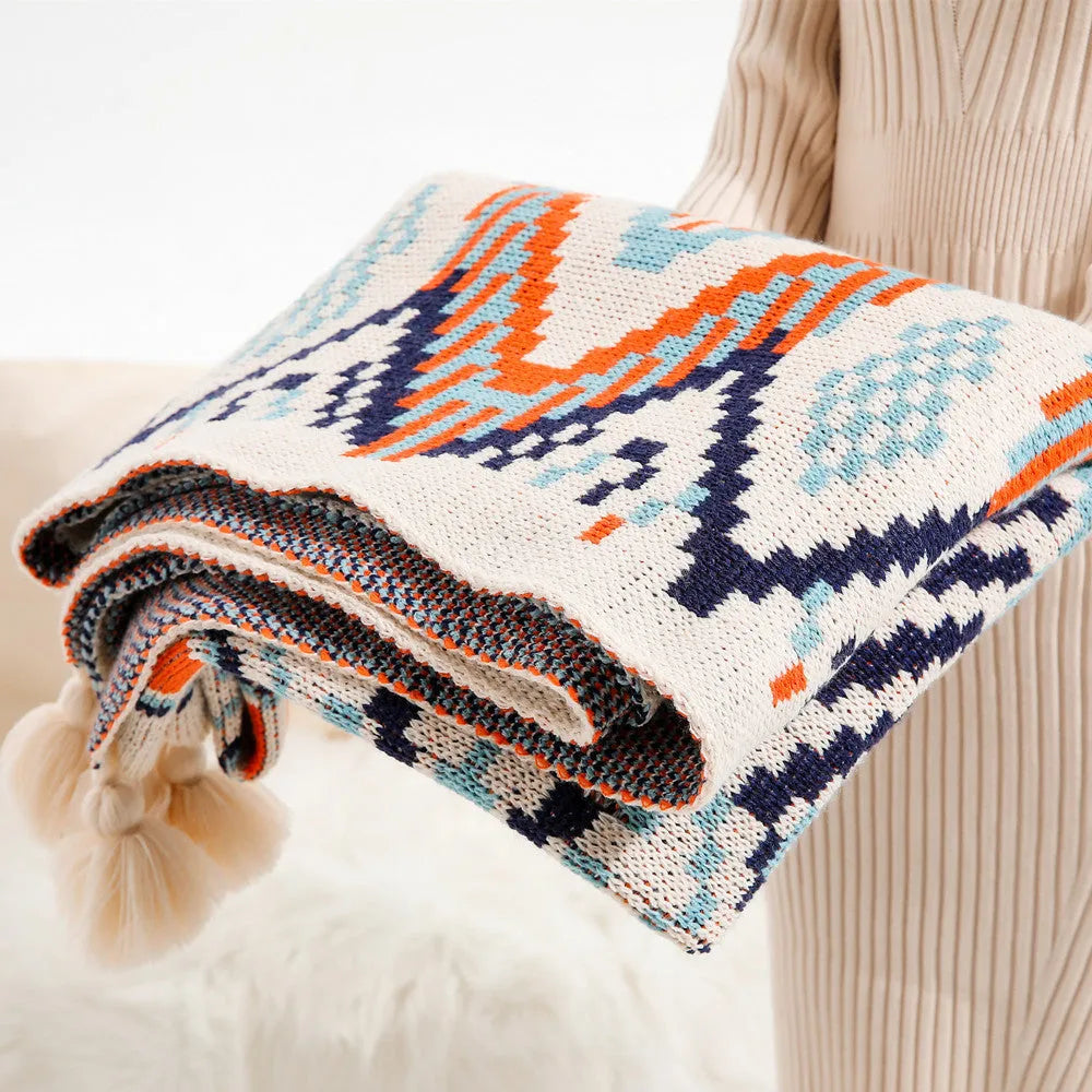 Bohemian style acrylic sofa blanket with tassel fringe in a geometric blue and pink pattern draped over a grey sofa.