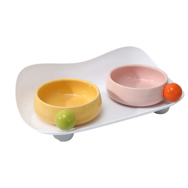 A pink ceramic raised pet bowl with two compartments and a non-slip black rubber base.