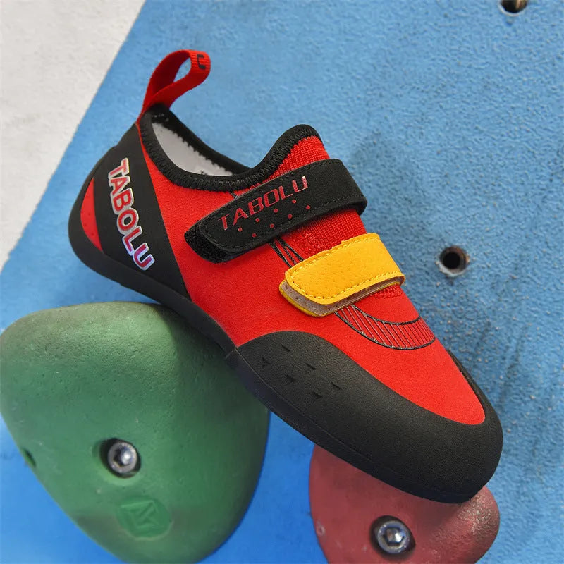 Beginner rock climbing shoes with Velcro strap in black and red colour scheme on a rocky surface.