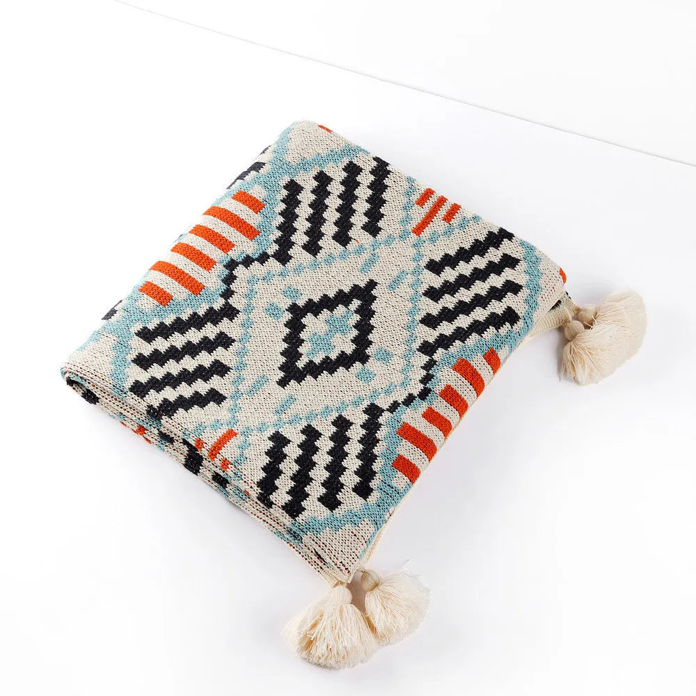 Bohemian style acrylic sofa blanket with tassel fringe in a geometric blue and pink pattern draped over a grey sofa.